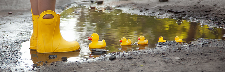 Billede af børnefødder med gummistøvler på og en lille række af gummiænder i en vandpyt